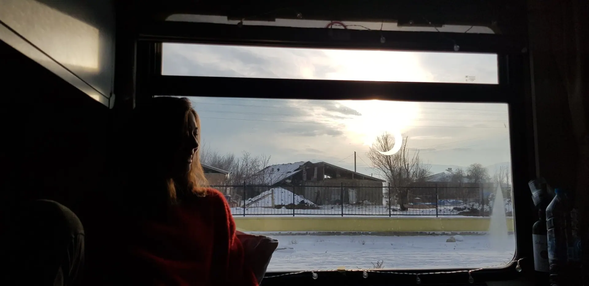 A woman in a red sweater sits in silhouette at a dark window, looking out at snow and a low winter sun