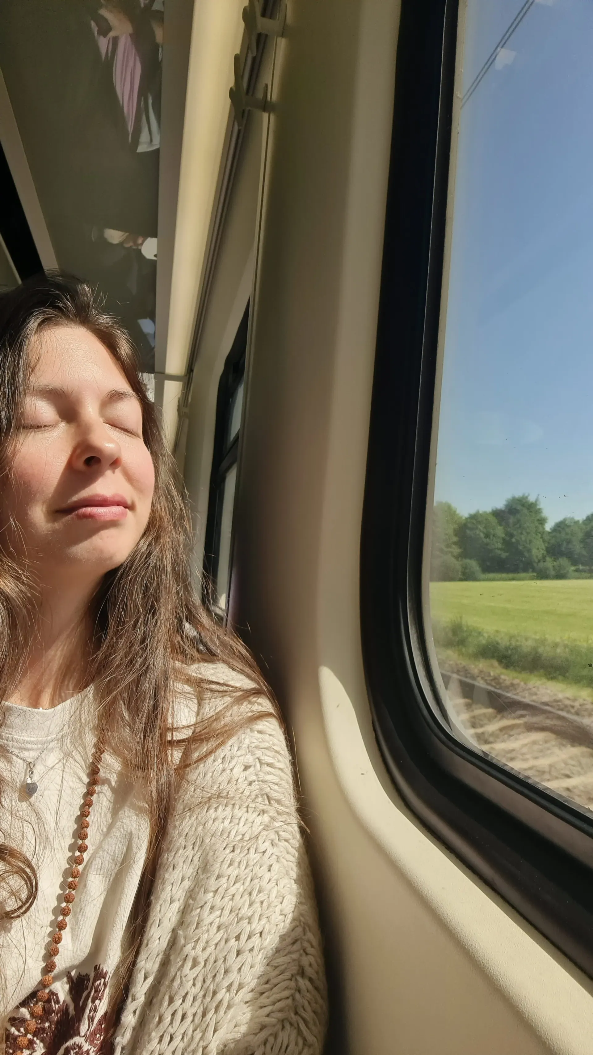 Ece resting against a train window with her eyes closed, green fields outside