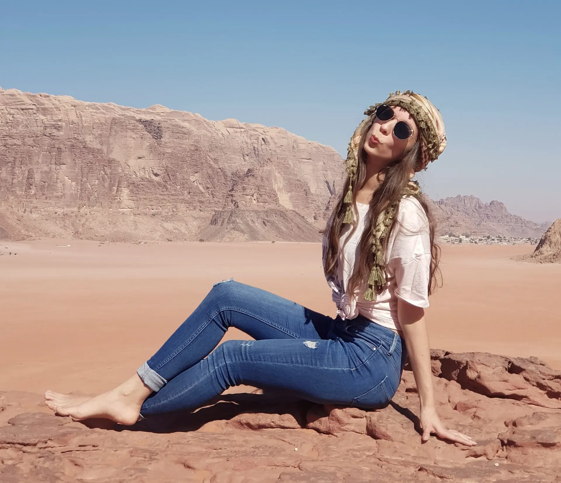 Ece sitting on red rock in a wide desert, in sunglasses and a tasselled headscarf, with sandstone cliffs behind her