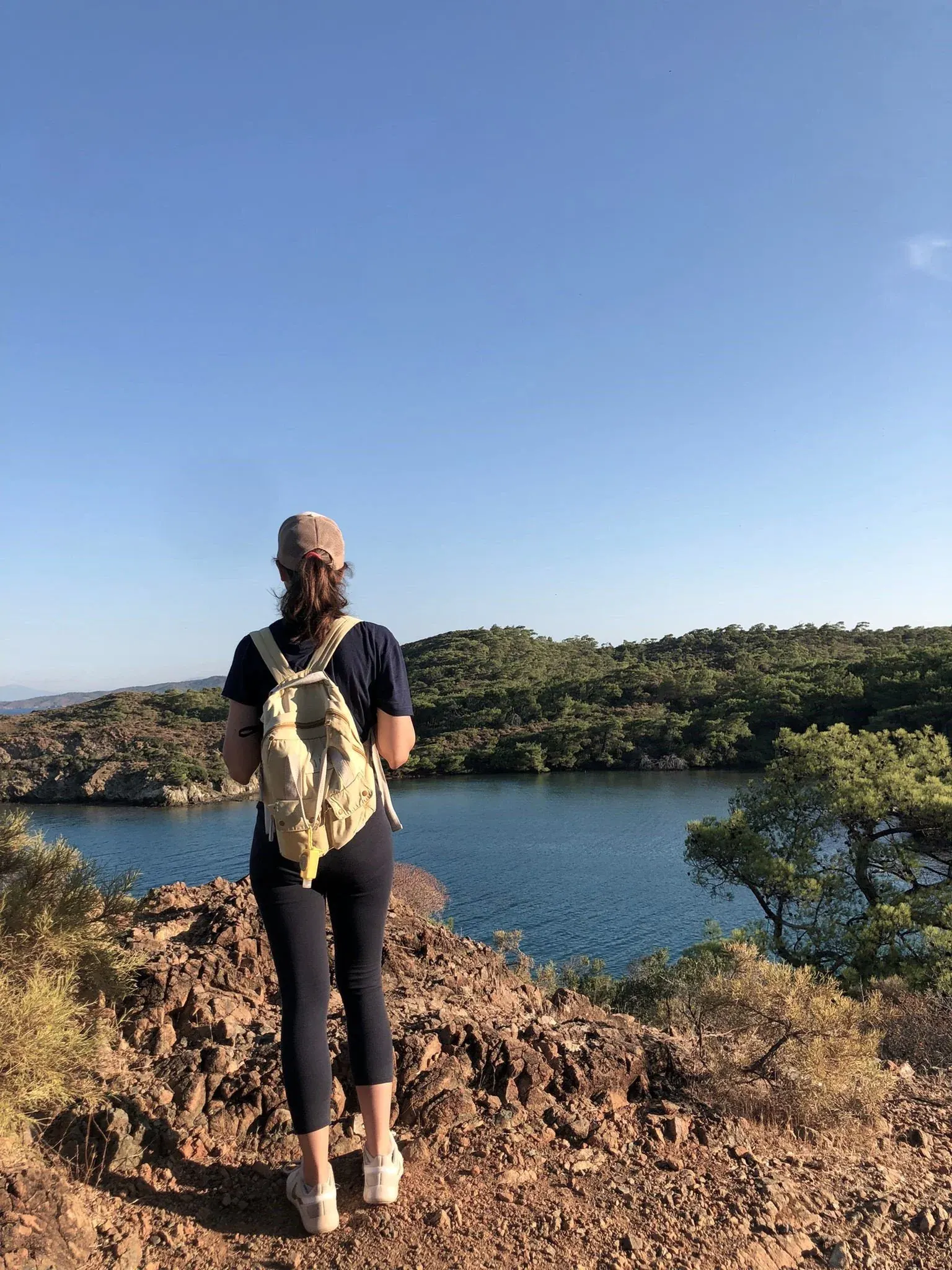A woman in a cap and backpack, seen from behind, looking out over a bay and green hills