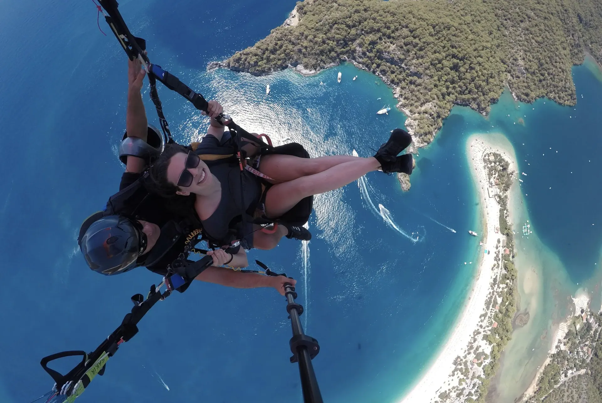 A woman smiling on a tandem paraglide, high above a turquoise lagoon and a white sand spit