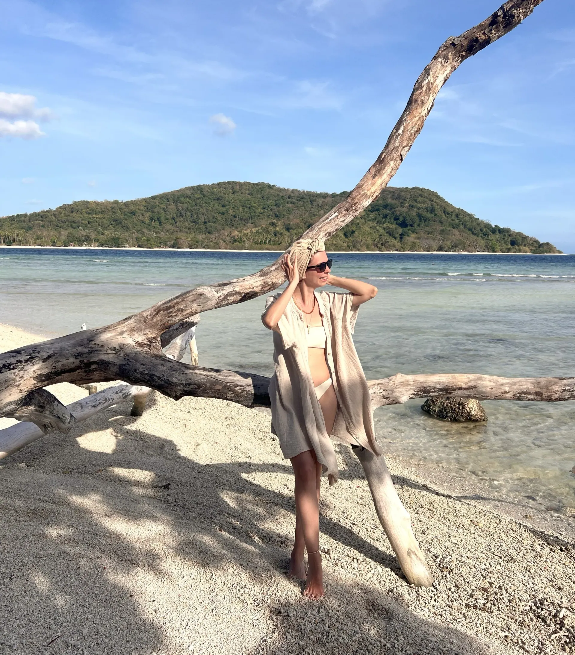 Ece standing by a driftwood tree on a beach, an island behind her
