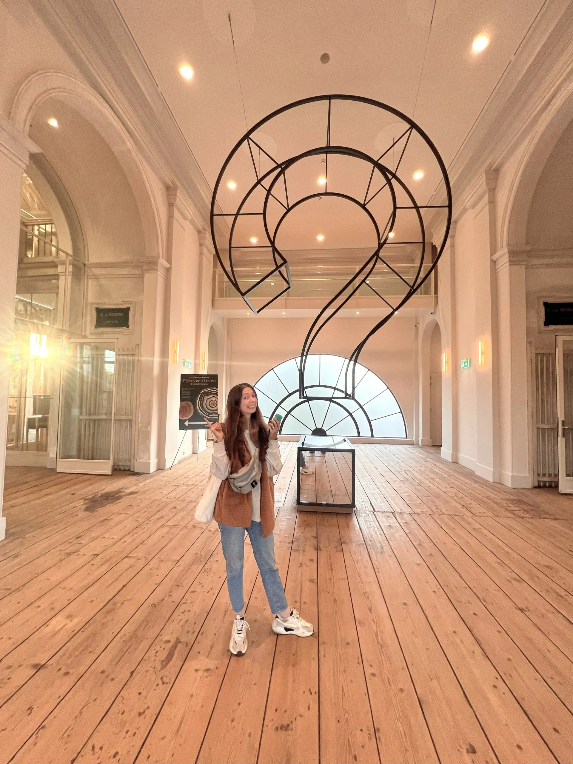 Ece standing beneath a large question-mark sculpture in a bright hall