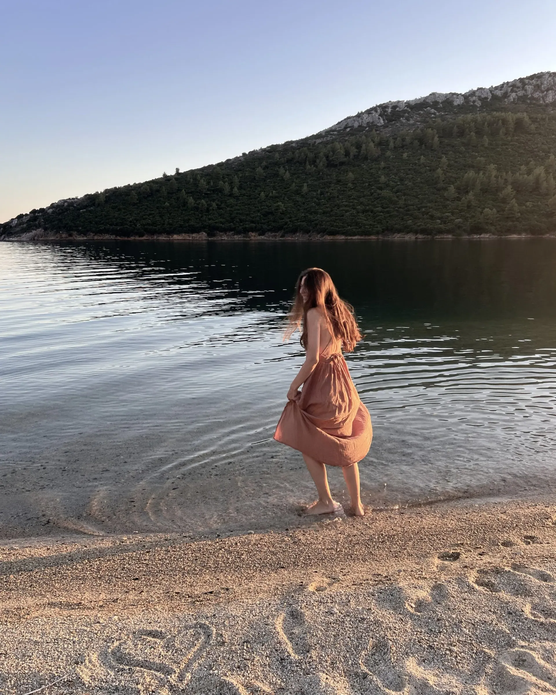 Ece in a long dress stepping into calm water at the shore