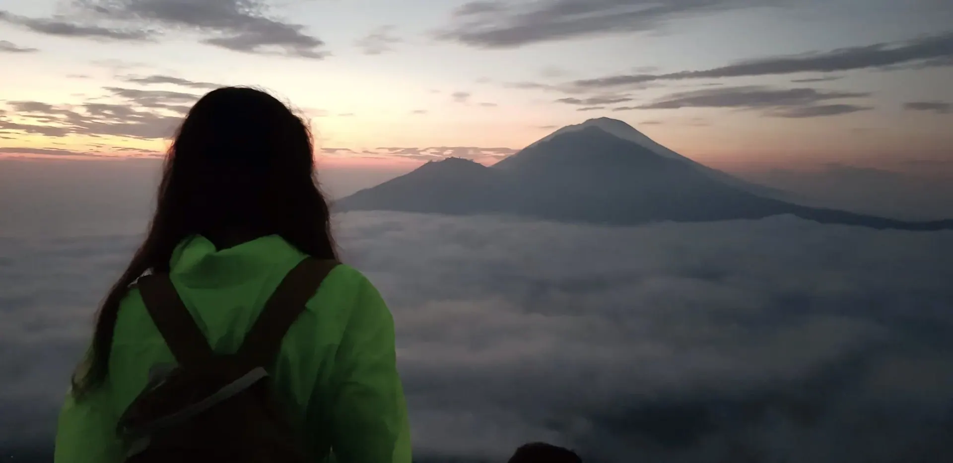 A woman in a bright green jacket and backpack, seen from behind, looking out at a mountain peak above a sea of clouds
