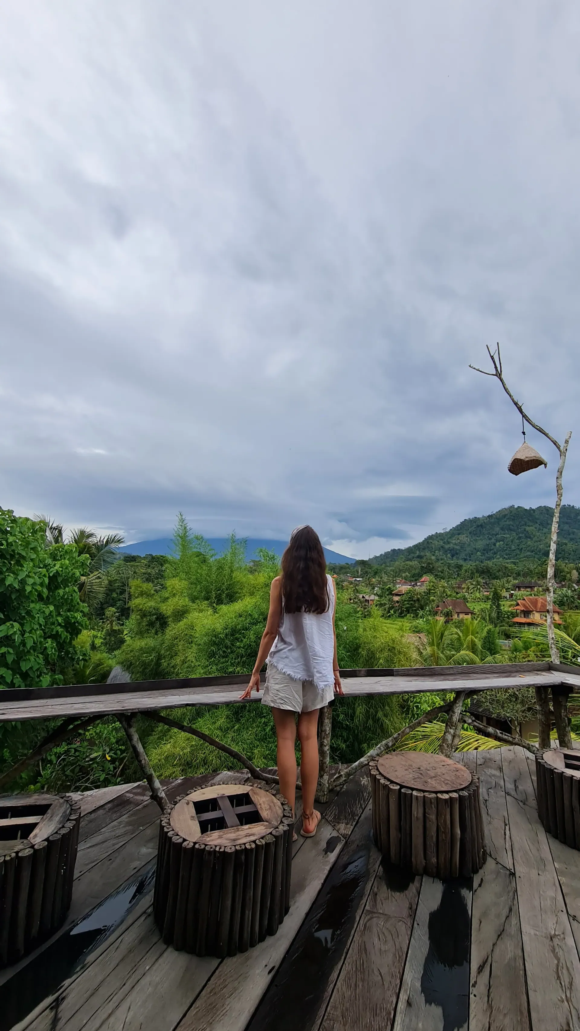 A woman seen from behind on a wooden deck, looking out over green tropical hills and a cloud-covered mountain