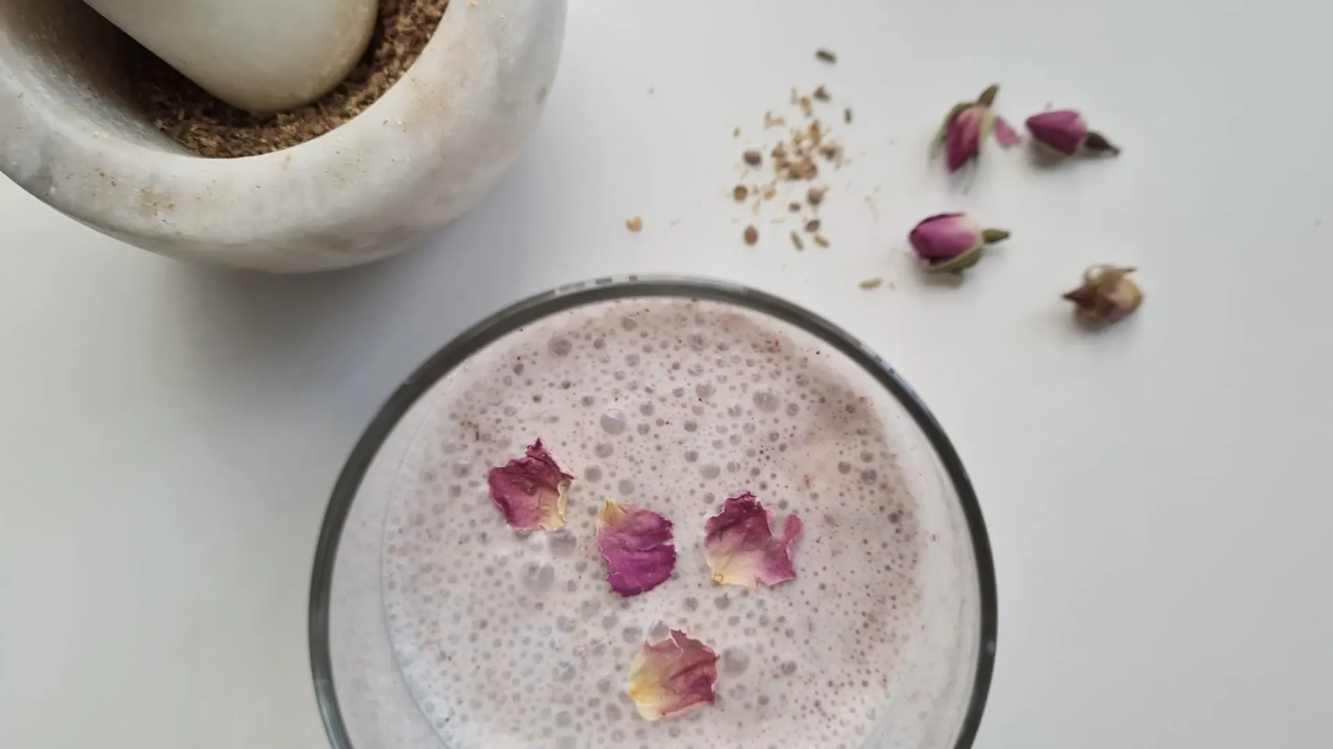 A glass of frothy pink drink topped with rose petals, beside a mortar of ground spices and dried rosebuds