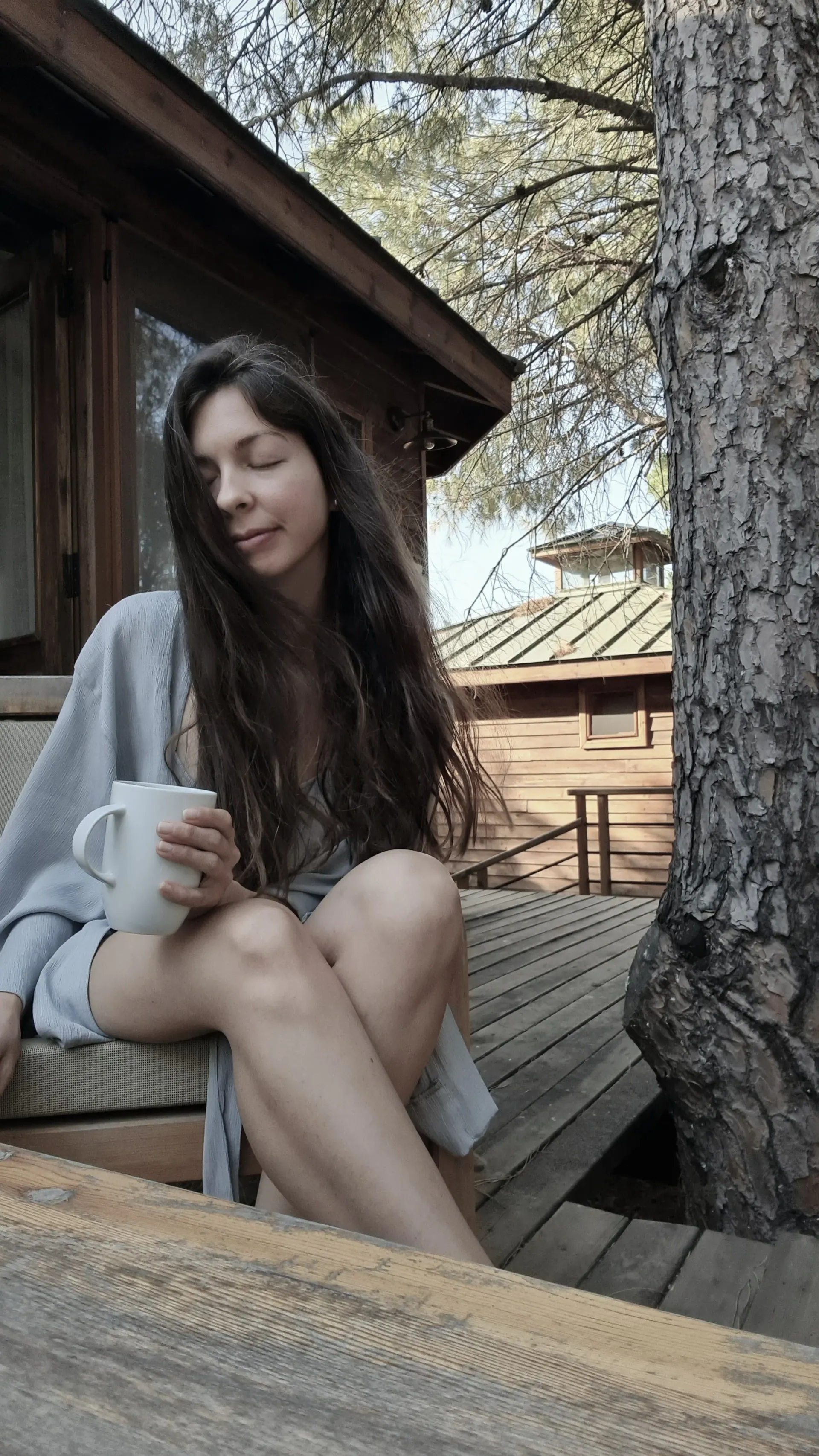 A woman sitting on a wooden deck beside a pine tree, eyes closed, holding a white mug
