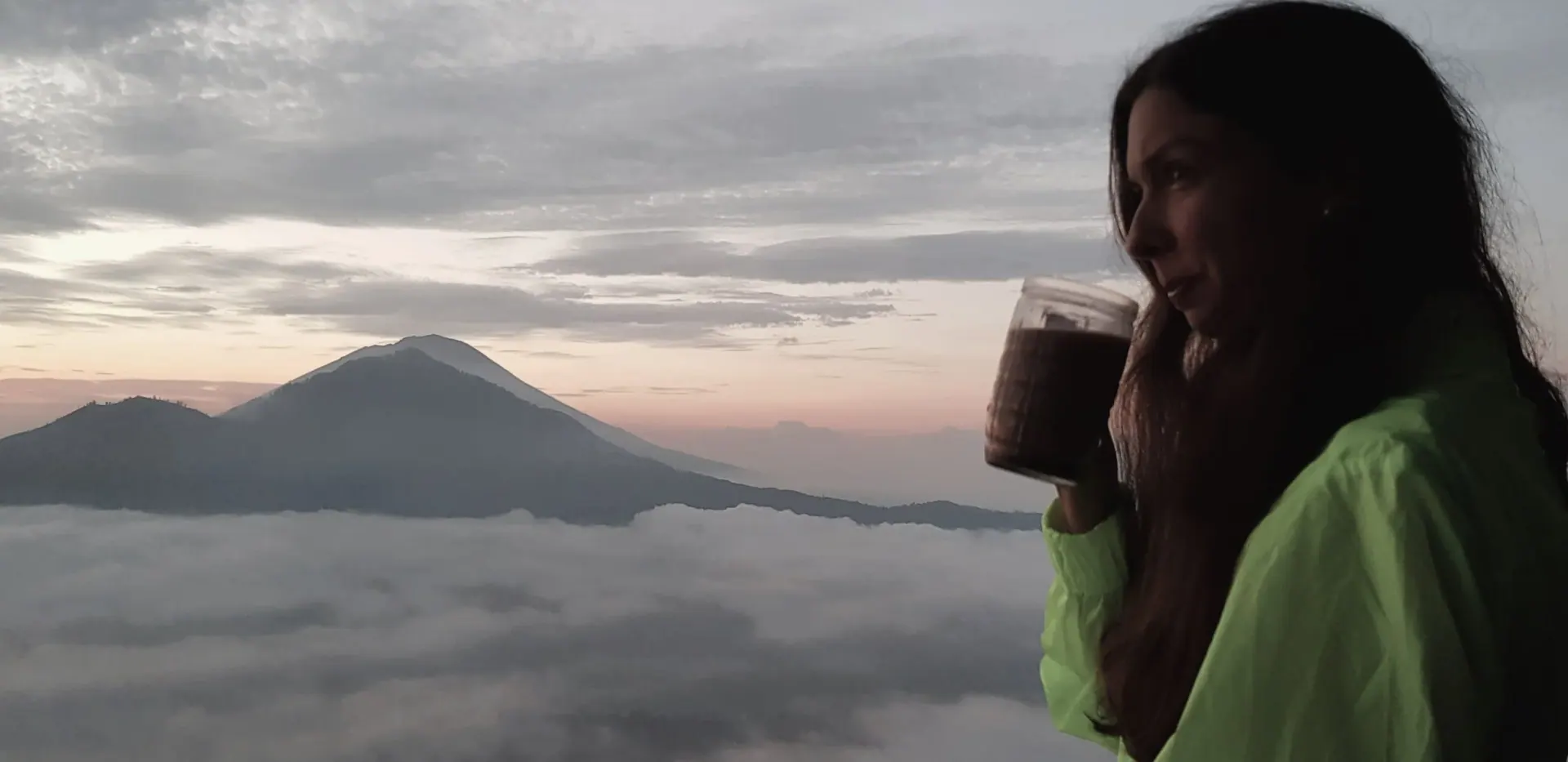 A woman in a bright green jacket sipping from a glass mug, with a mountain peak rising above a sea of clouds in the distance