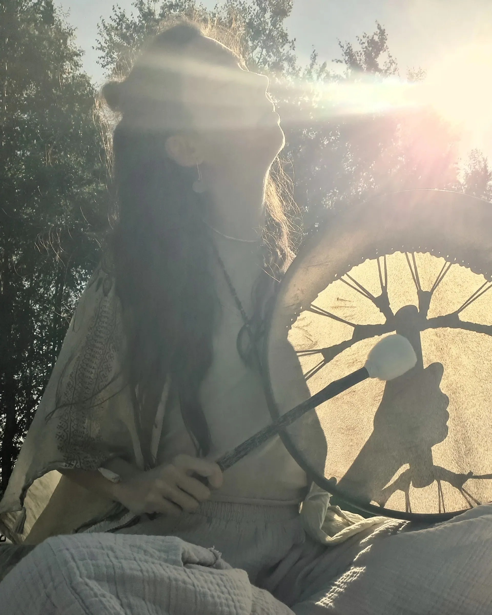 A woman with long hair playing a frame drum, her face washed out by bright sunlight, trees behind her
