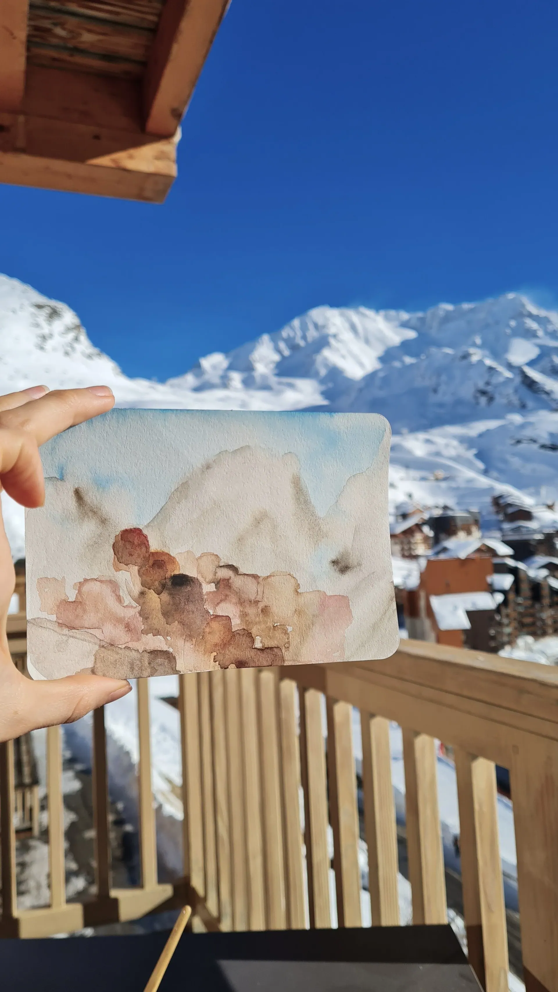 A hand holding a small watercolour of mountains in front of a snowy mountain view