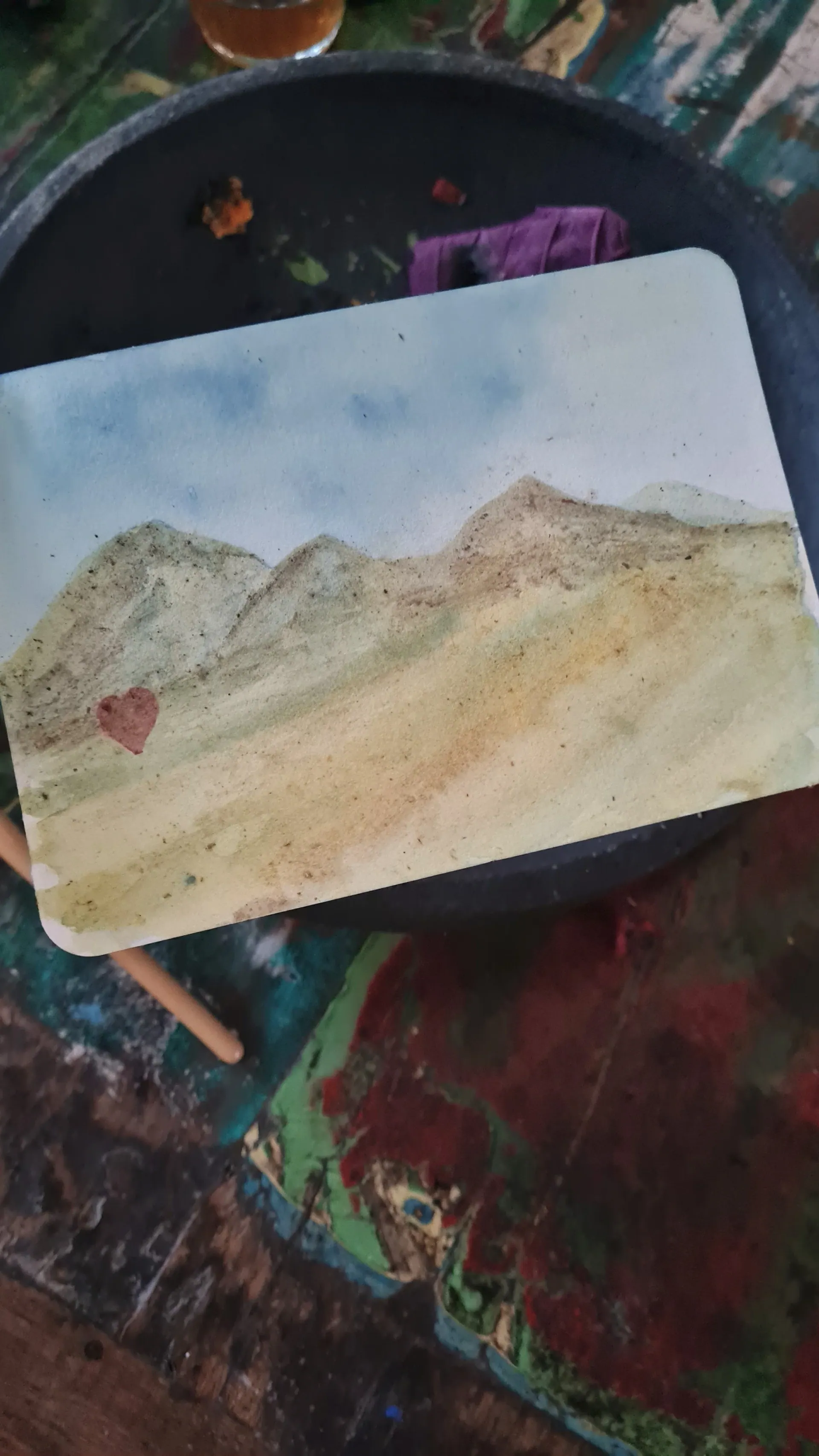 A small watercolour of green and golden mountains with a little red heart, resting on a stone mortar on a colourful table