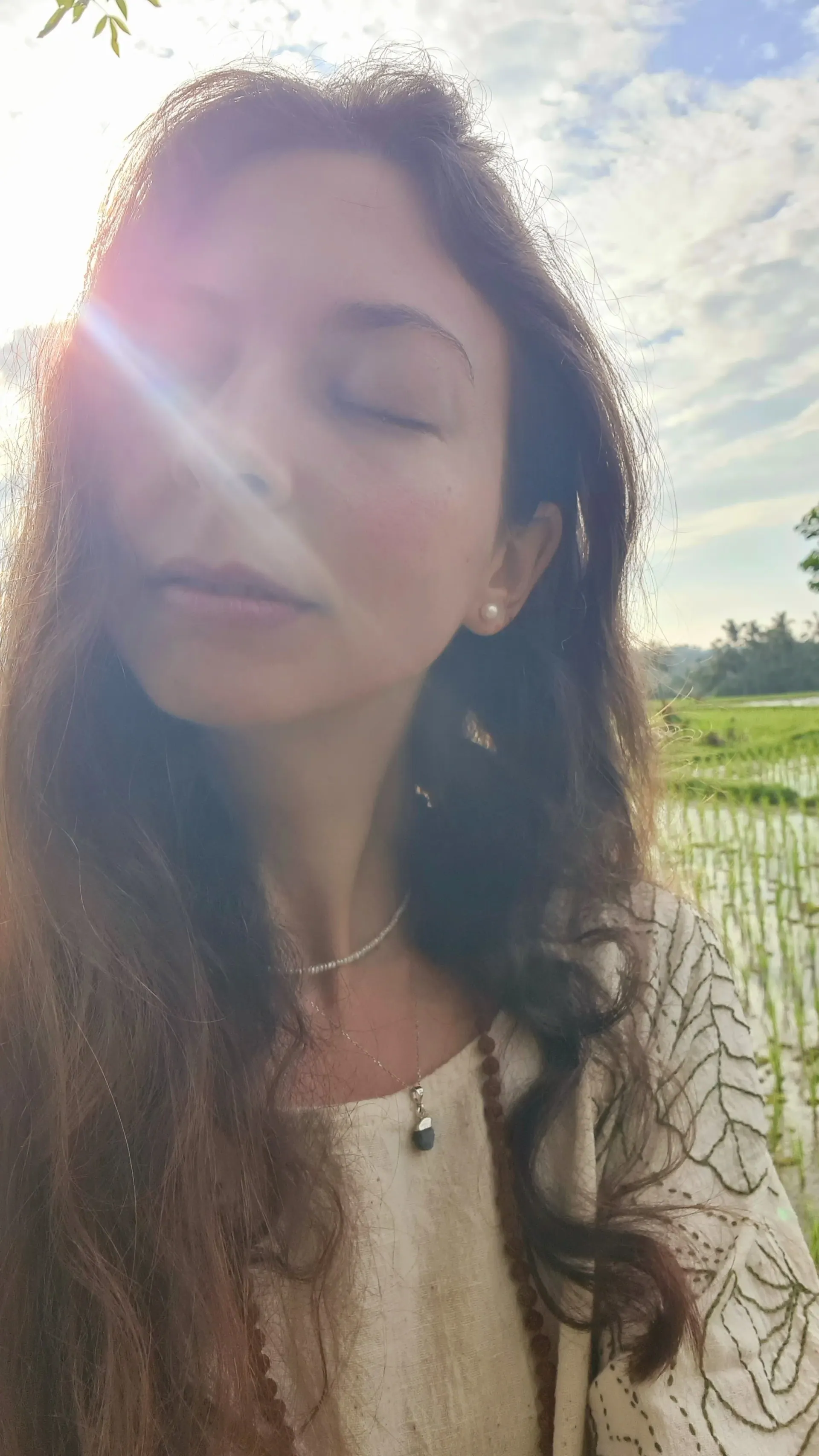A woman with long wavy hair and closed eyes, sunlight flaring across her face, with a green rice field behind her