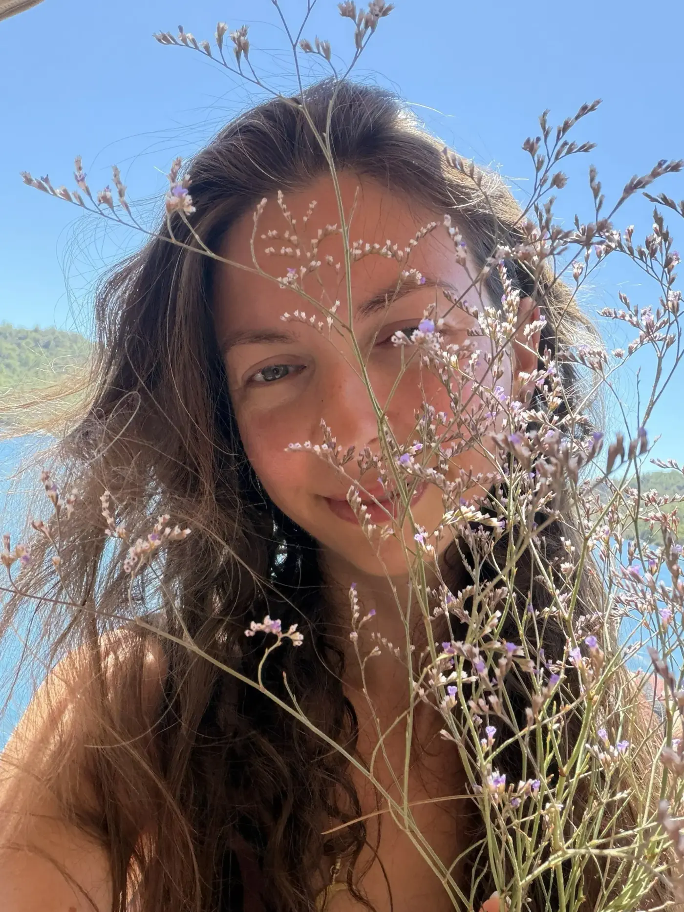Ece smiling softly through sprigs of tiny lilac flowers, with the sea and a blue sky behind her