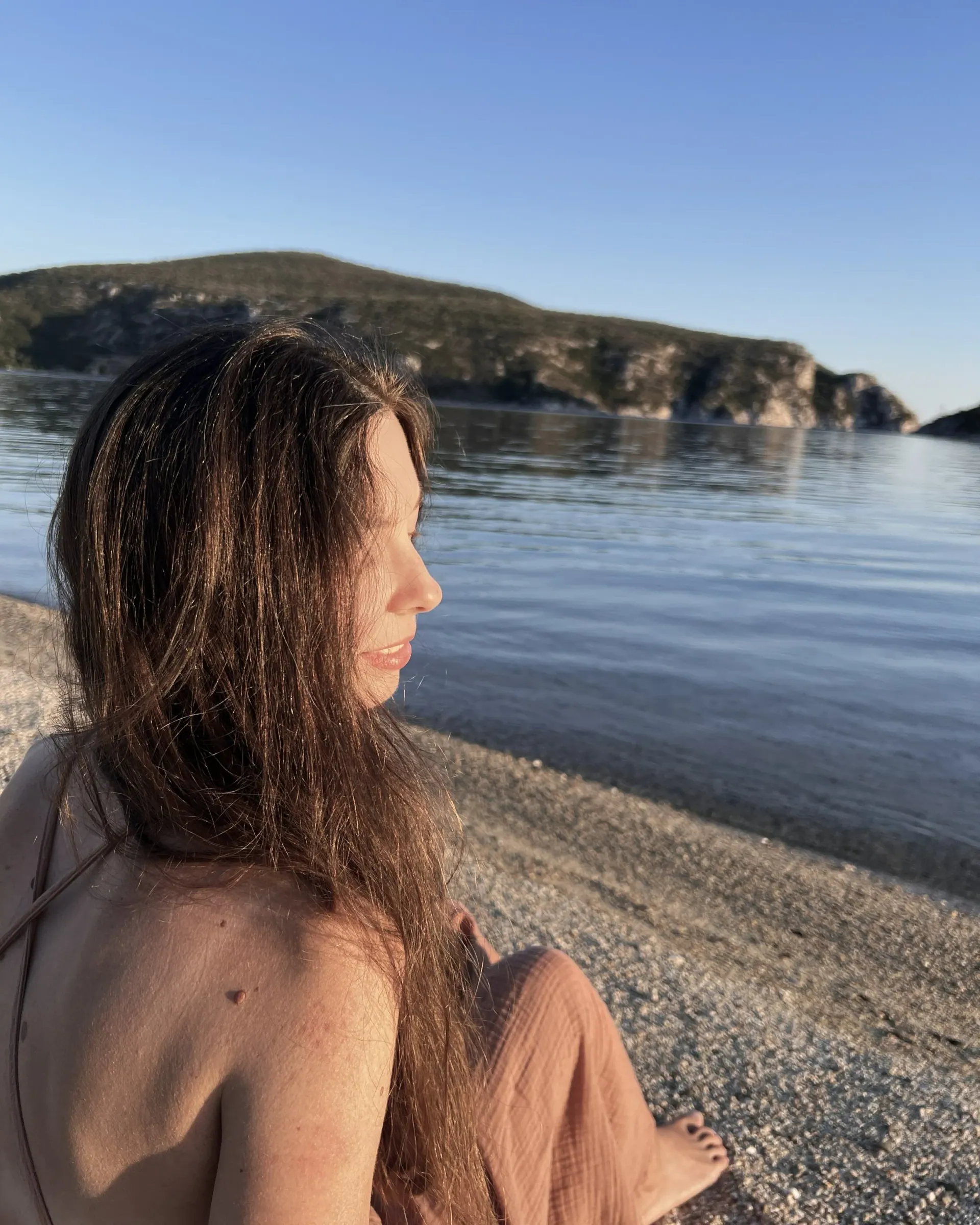 Ece with long hair sitting on a pebble beach, looking out over the sea