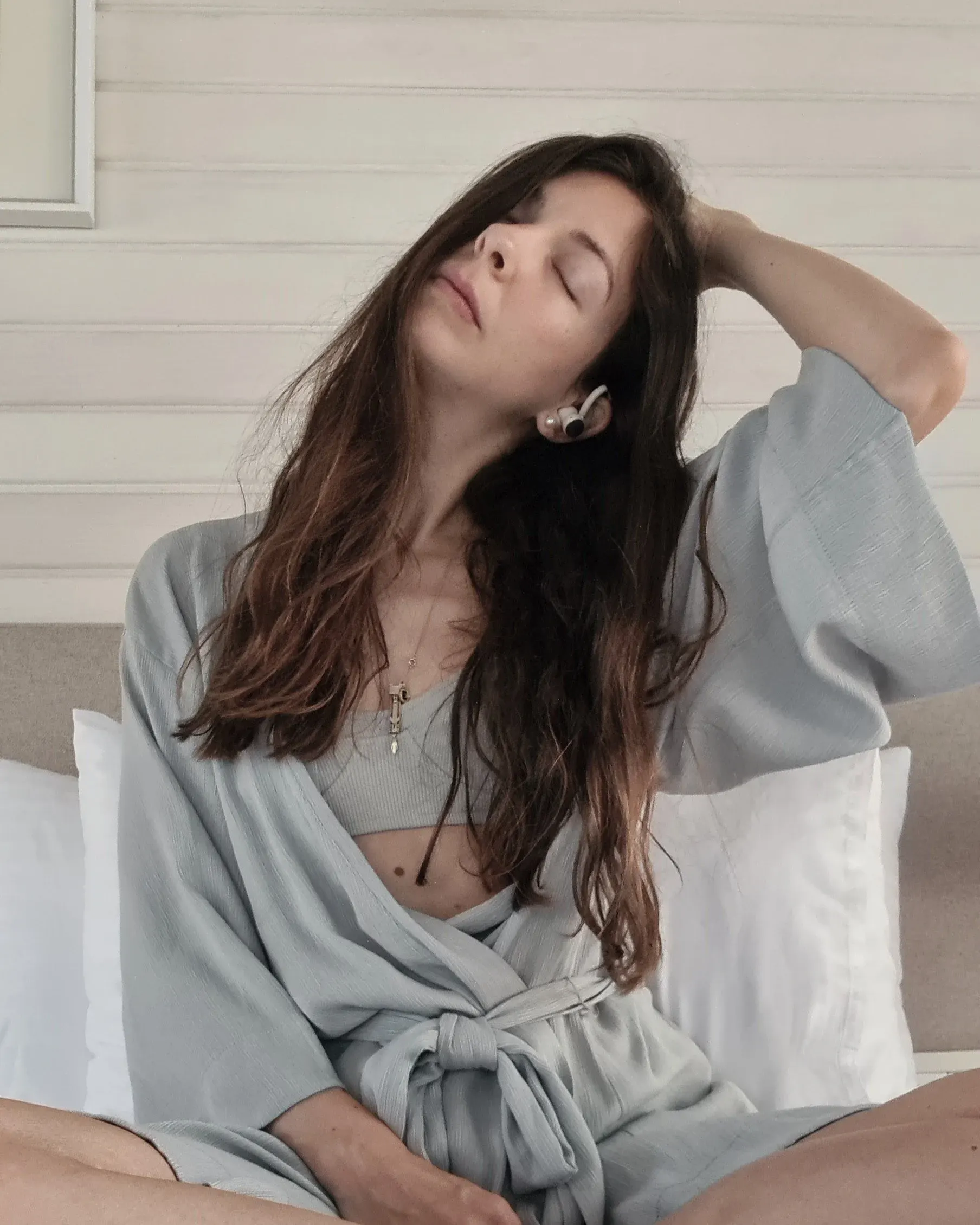 Ece in a soft grey-blue robe on a bed with white pillows, eyes closed, one hand in her hair as she stretches her neck