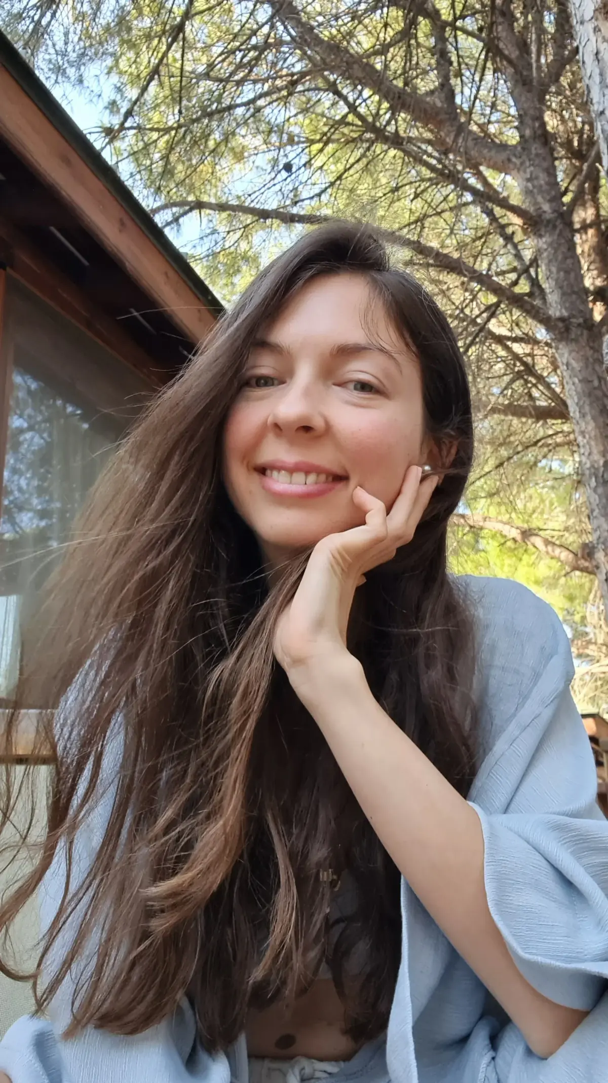 Ece smiling with her chin resting on her hand, outdoors under pine trees