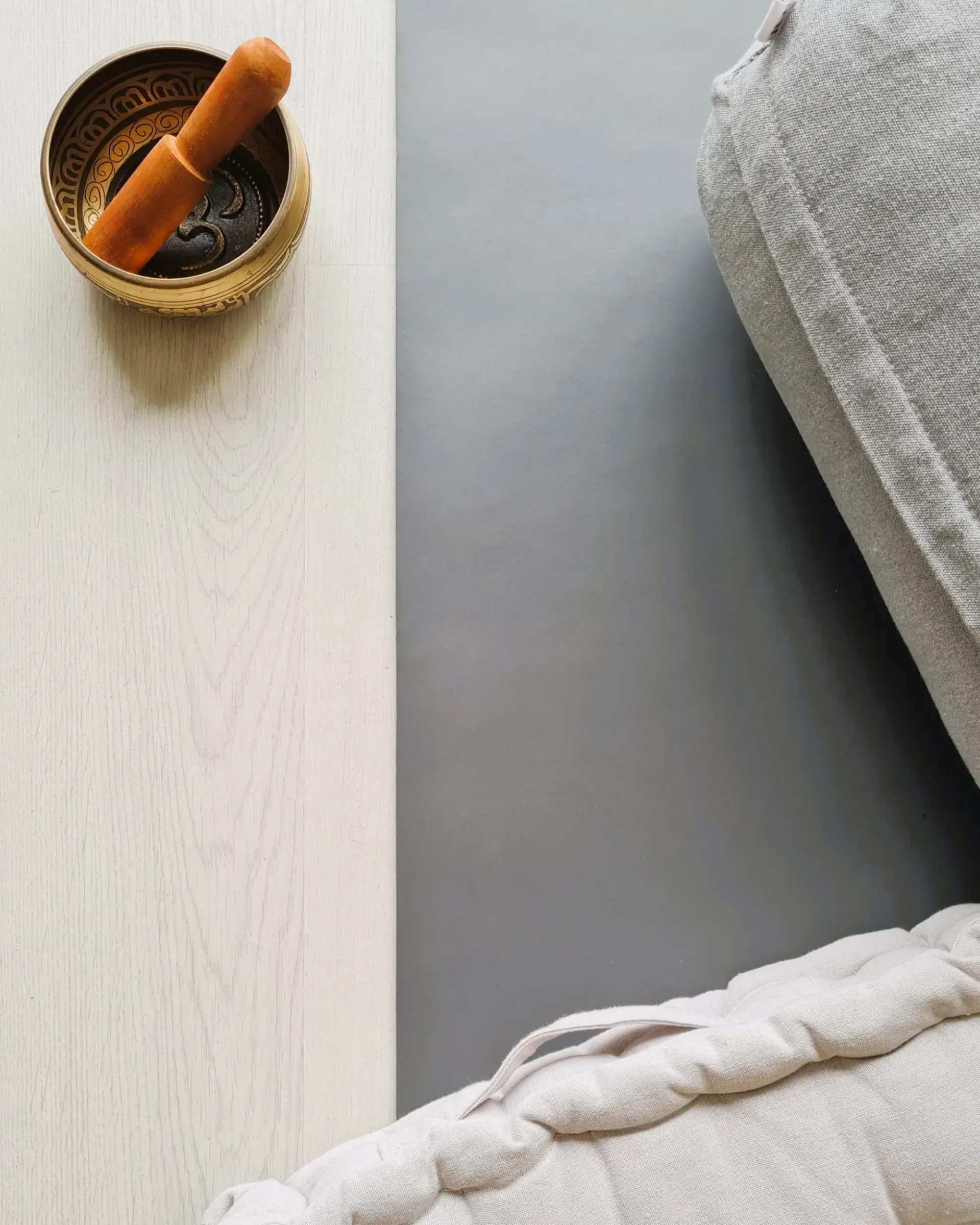 A brass singing bowl with a wooden mallet on a pale wooden floor, beside a grey mat and soft cushions